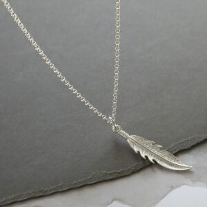 A delicate silver feather pendant hanging from a silver belcher chain over a grey slate