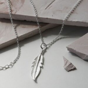 Delicate silver feather hung on a silver belcher chain laying over light brick coloured stone