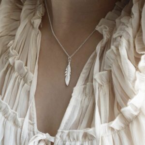 A delicate silver feather pendant hanging from a silver belcher chain round a woman's neck surrounded by white see through material