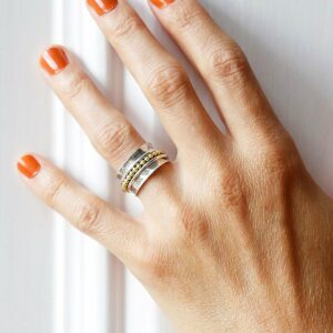 Self care spinning ring with gold detail on model with Orange nail polish against a white wall