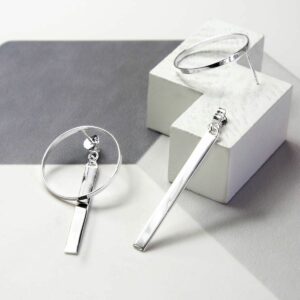 Large circular silver earrings with a long silver bar dangling from the top of them with a grey background laying on white blocks