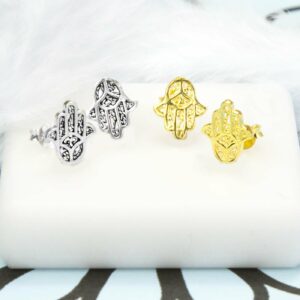 Pair of Silver Hamsa Hand Stud next to a Pair of Gold Hamsa Hand Studs on white tile background and blue and black pattern paper