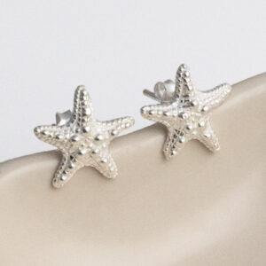 Delicate sterling silver stud earrings shaped like starfish, with fine textured detailing that captures the natural beauty of ocean life on a cream shall background.