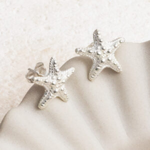 Delicate sterling silver stud earrings shaped like starfish, with fine textured detailing that captures the natural beauty of ocean life on a cream shall background.