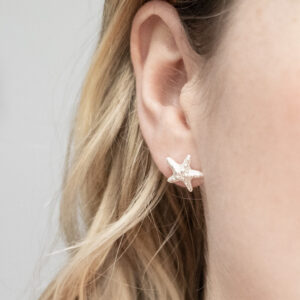 Delicate sterling silver stud earrings shaped like starfish, with fine textured detailing that captures the natural beauty of ocean life worn by a model with blonde curly hair.