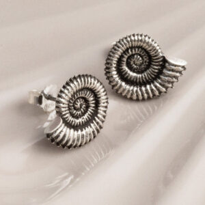 Sterling silver stud earrings featuring fossil-inspired designs with an oxidised finish, highlighting the intricate spiral patterns. A unique, nature-inspired accessory with a vintage, textured look on a cream shell background.