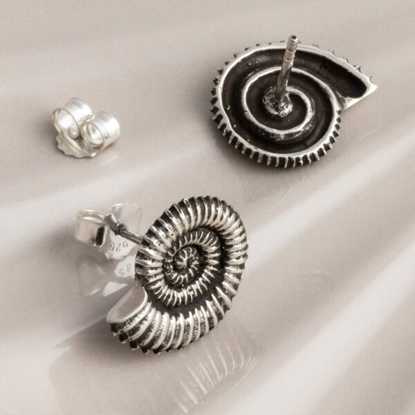 Sterling silver stud earrings featuring fossil-inspired designs with an oxidised finish, highlighting the intricate spiral patterns. A unique, nature-inspired accessory with a vintage, textured look on a cream shell background.