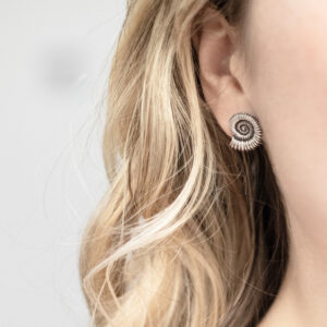 Sterling silver stud earrings featuring fossil-inspired designs with an oxidised finish, highlighting the intricate spiral patterns. A unique, nature-inspired accessory with a vintage, textured look worn by a model with curly blonde hair.