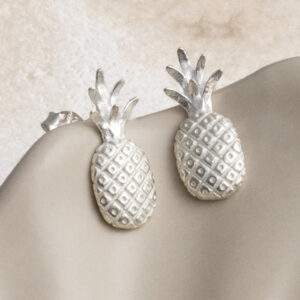 Playful sterling silver stud earrings shaped like pineapples, featuring fine detailing for a tropical, summery touch on a cream wavy cup background.