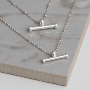 Silver sterling silver T-bar necklace on a grey tile background, showing fine gold sterling silver ball chain and small and large horizontal bar pendant.