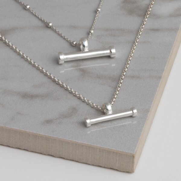 Silver sterling silver T-bar necklace on a grey tile background, showing fine gold sterling silver ball chain and small and large horizontal bar pendant.